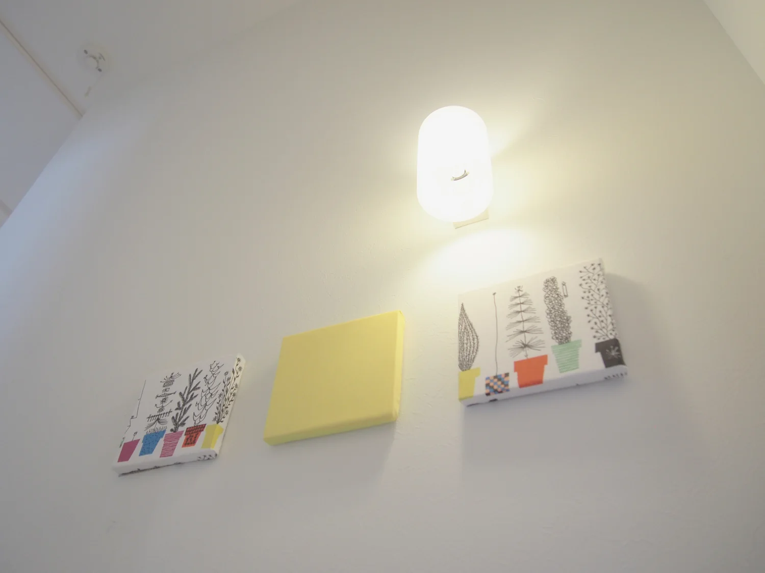 壁面の照明と3枚のファブリックパネル。真ん中は黄色いパネルで、両側の二枚は観葉植物が描かれている。